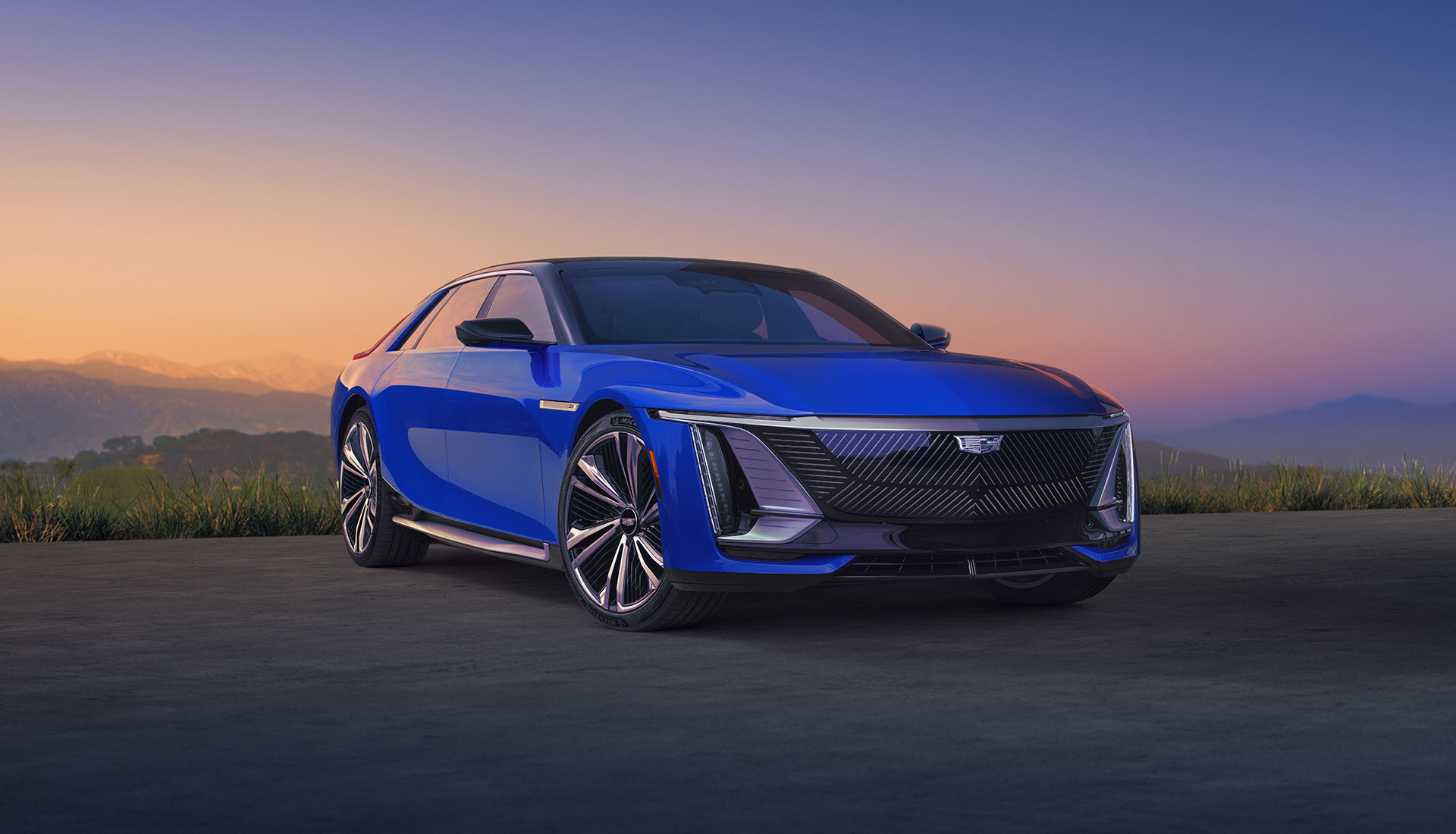 Front three-quarter view of a blue 2026 Cadillac Celestiq parked on a scenic road at sunset, with sleek LED headlights, large alloy wheels, and mountains silhouetted in the background.