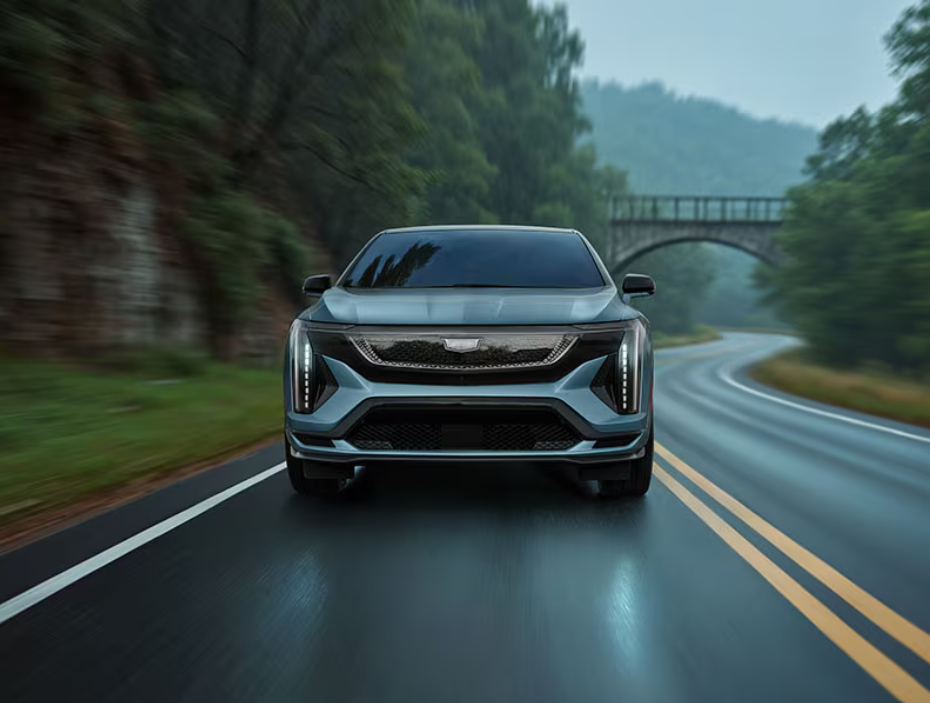 Front view of a silver 2026 Cadillac OPTIQ-V driving on a wet, winding road, with vertical LED daytime running lights, a sporty grille, and motion blur from surrounding trees and a bridge in the background.