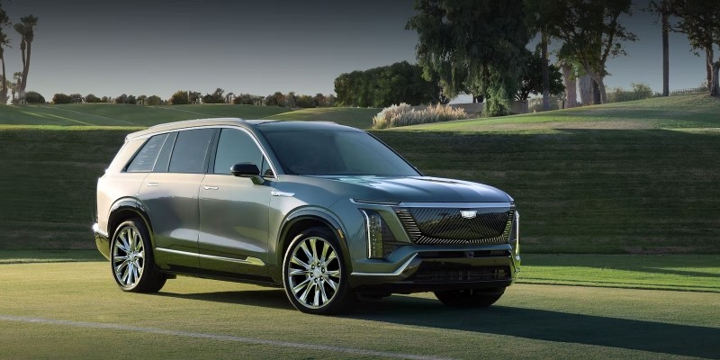 Front three-quarter view of a gray 2026 Cadillac VISTIQ parked on a manicured grassy landscape, featuring a bold illuminated grille, vertical LED headlights, large multi-spoke wheels, and trees in the background under soft daylight.